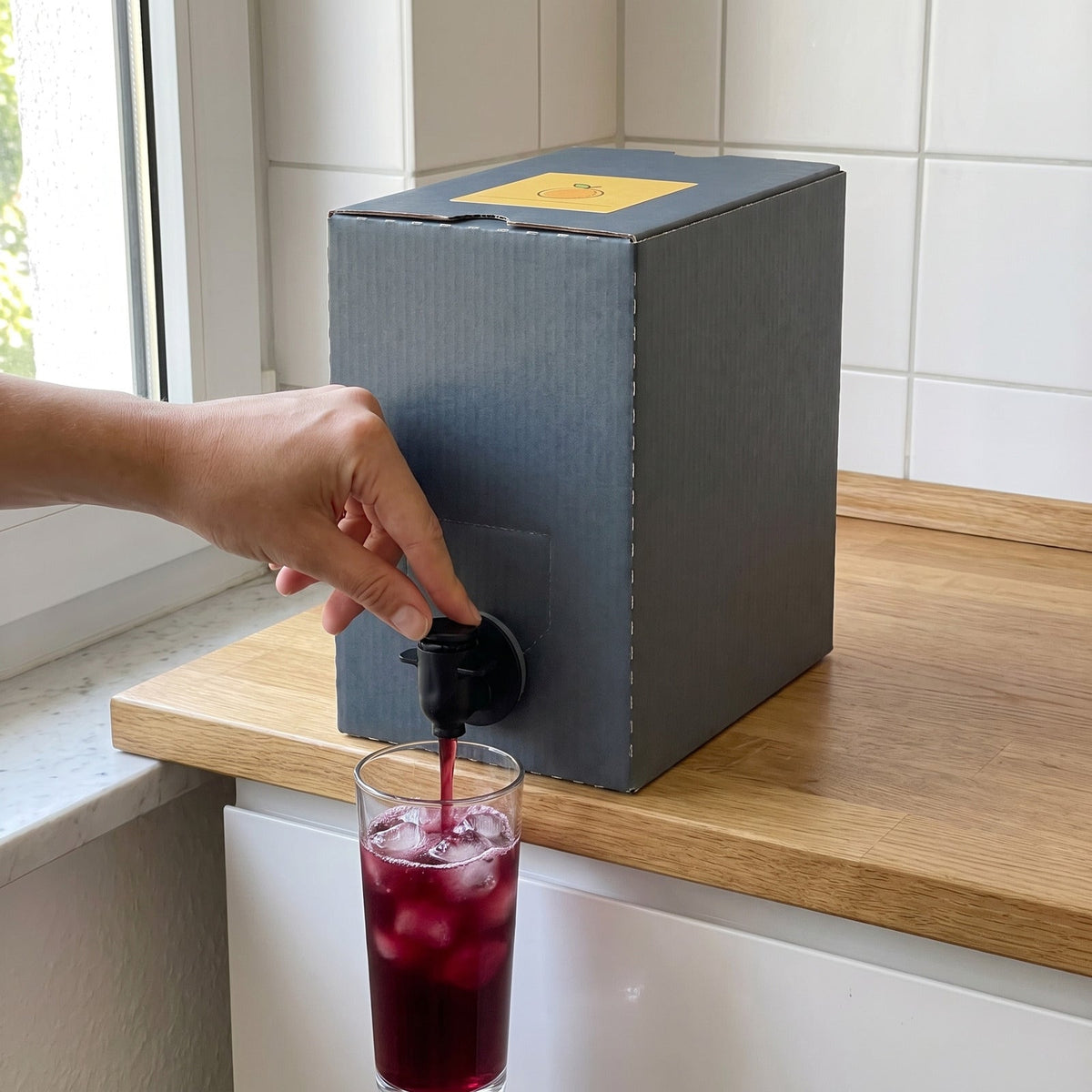 Hand zapft Getränk aus der VitaTap Box in ein Glas mit Eis auf der Küchenarbeitsplatte